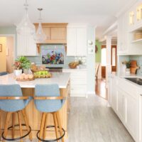 White kitchen cabinets and countertops, rift cut white oak range hood and kitchen island, color mosaic backsplash light blue fabric high-backed bar stools, kitchen sink