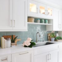 White kitchen cabinets with light green tile subway tile backsplash behind kitchen sink, color mugs on shelf above sink