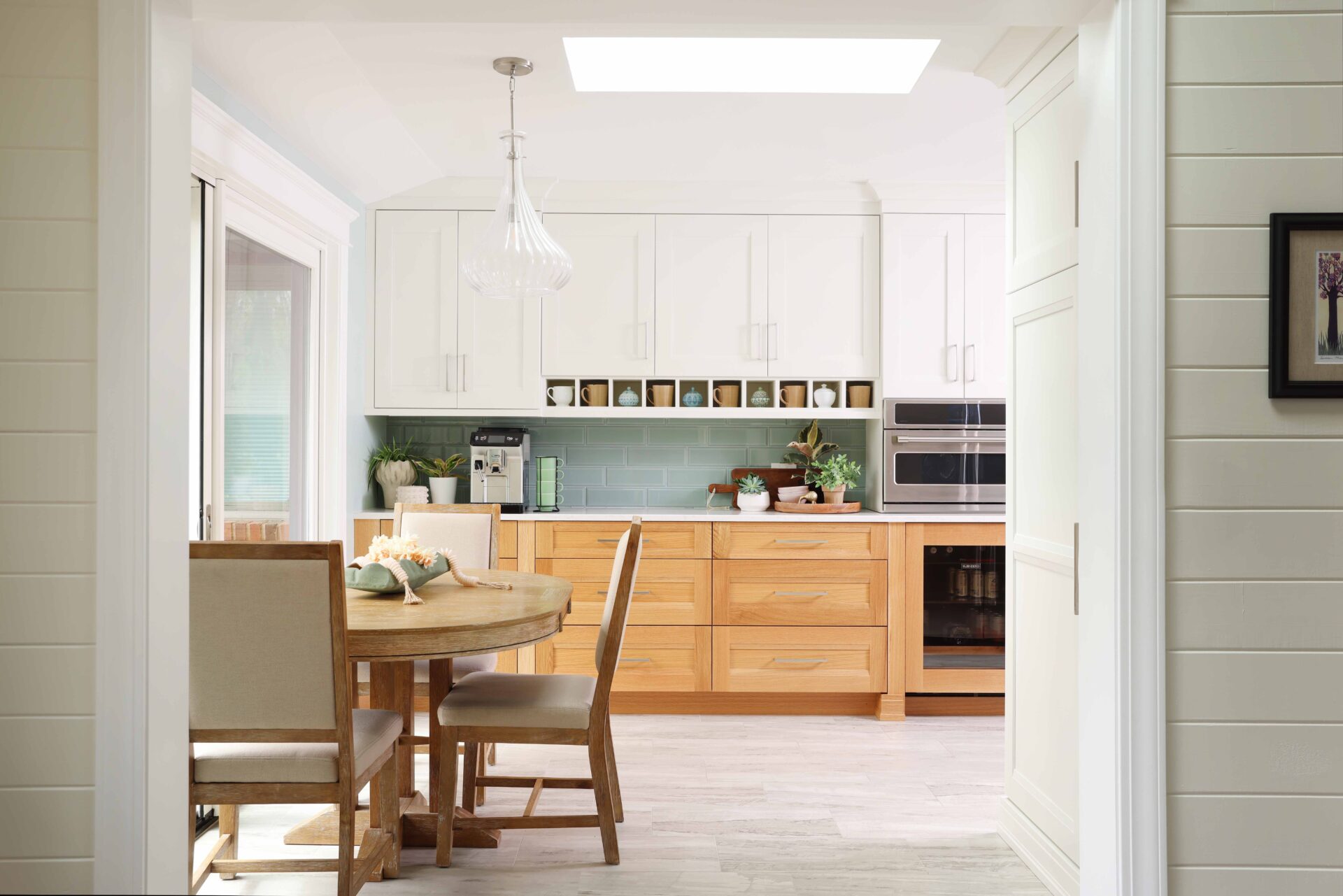 On the left, sliding patio door, and kitchen table with chairs. Coffee bar with upper white cabinets, rift cut white oak lower cabinets, coffee mugs in shelf nooks, light green tile backsplash, subway tile. Microwave and beverage/bar fridge refrigerator