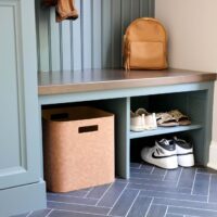 Mudroom with rainforest green cabinets, wood stained bench with bead board wainscot paneling detailing above. Shelving below bench. Lower shelves hold shoes and a brown storage bin. Herringbone gray tile floor tiles with white grout