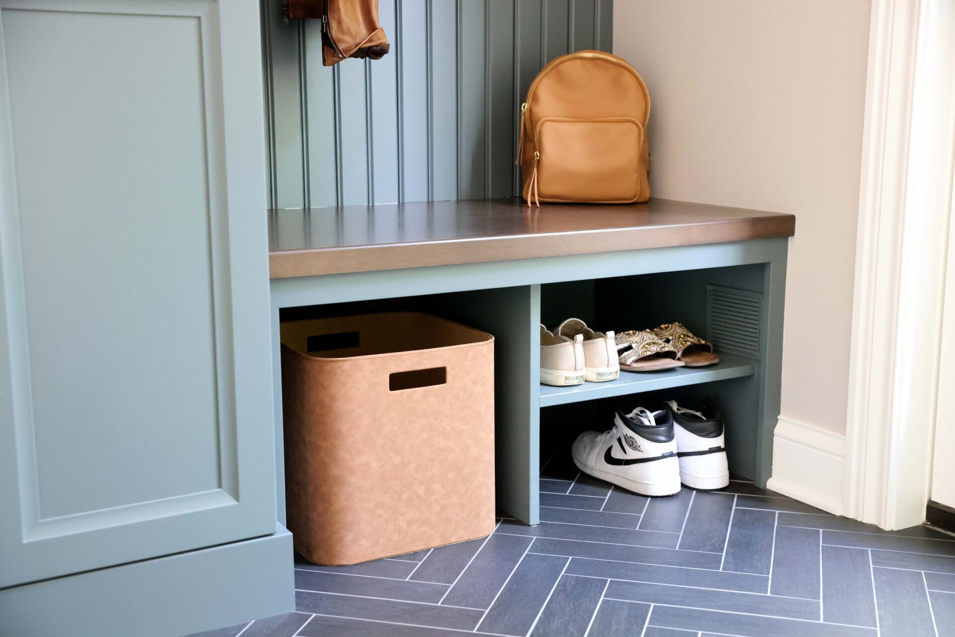 Mudroom with rainforest green cabinets, wood stained bench with bead board wainscot paneling detailing above. Shelving below bench. Lower shelves hold shoes and a brown storage bin. Herringbone gray tile floor tiles with white grout