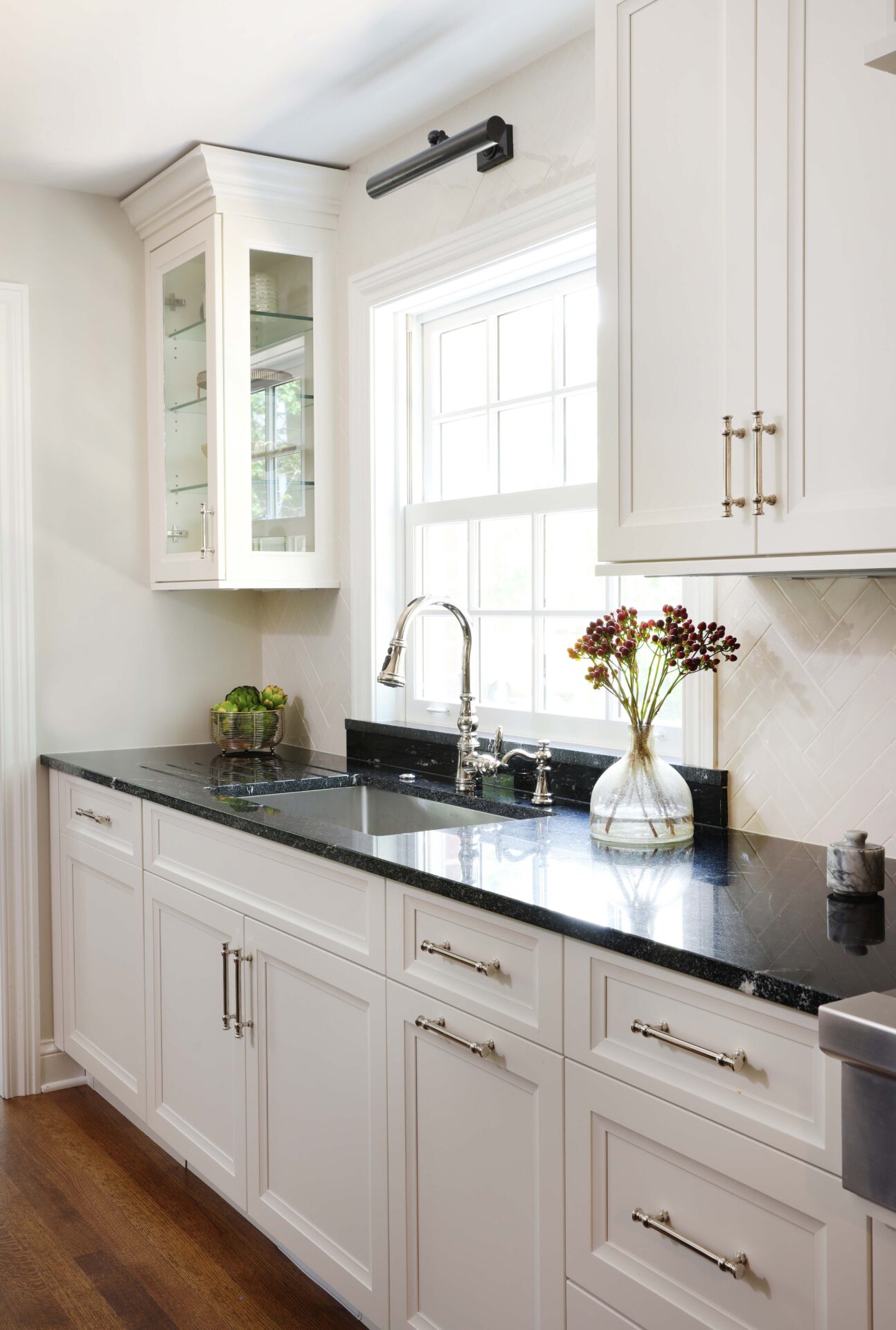 Polished nickel kitchen faucet and cabinet hardware, white kitchen cabinets, black countertops, one upper cabinet glass-fronted cabinet