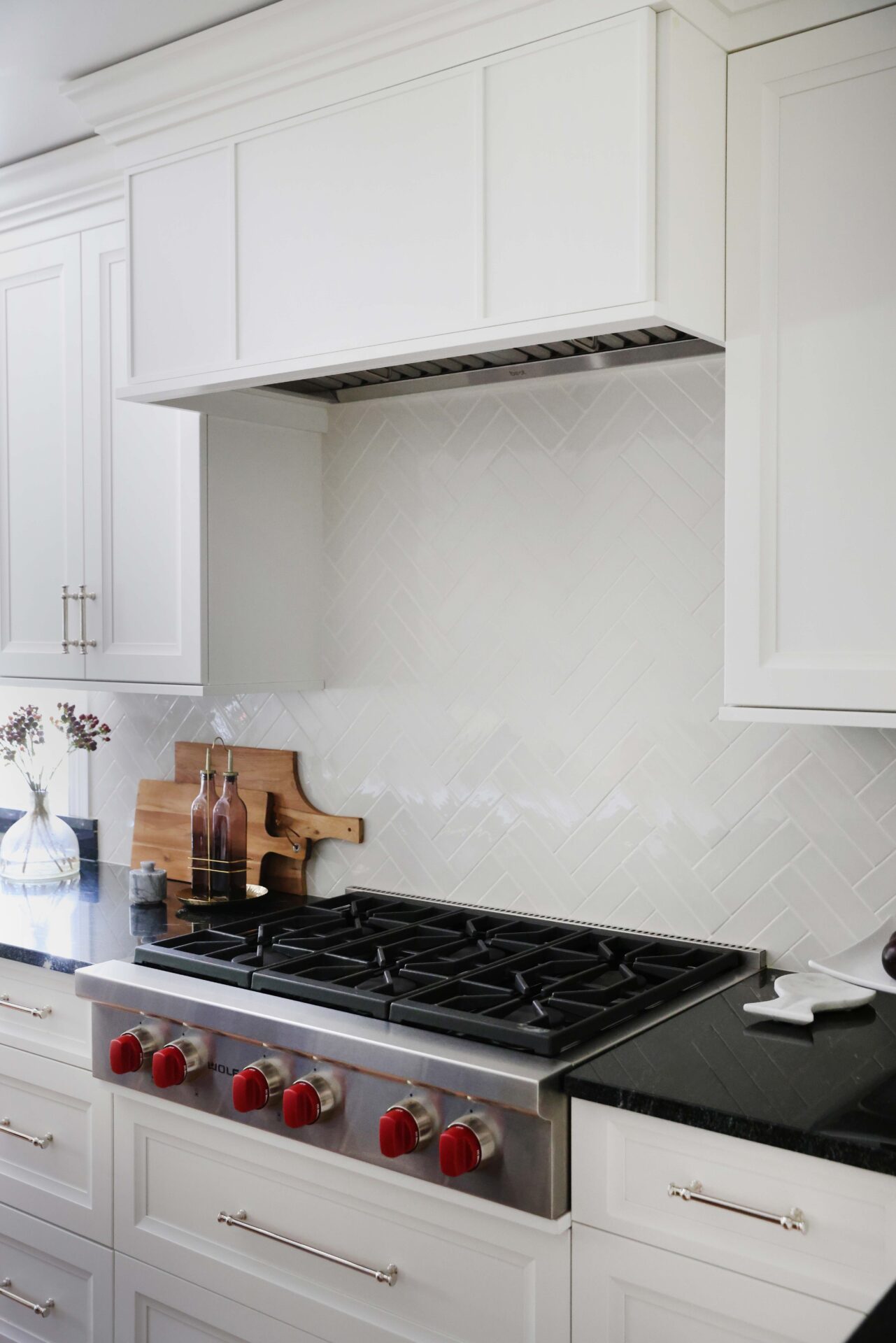 White herringbone backplash tile in kitchen, white range hood, range top with red knobs, white cabinets