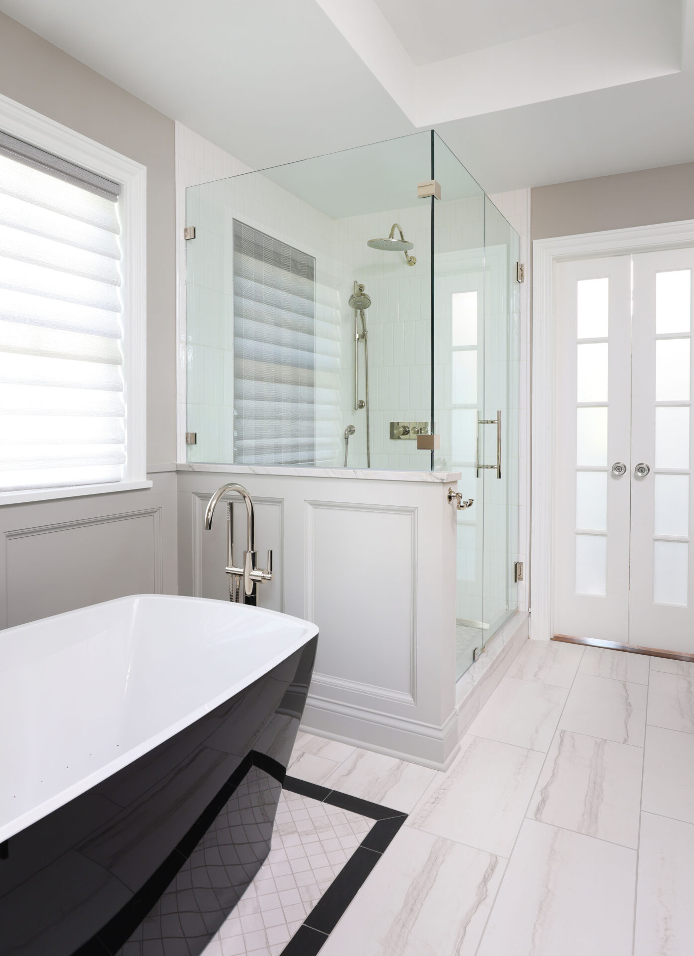 Freestanding tub with black exterior, white interior, polished chrome floor mounted faucet, white and black floor tile. Wainscoting half wall, glass shower with polished nickel rain shower head