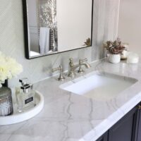 Polished nickel faucet, black rimmed mirror white quartzite countertops thick edge. Honey bronze knobs, silver textured tile vanity wall
