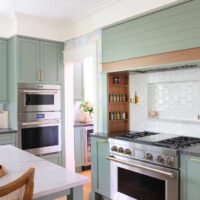 Natural stone white countertop at kitchen island, stacked stainless steel oven and microwave, light green cabinets, green hood with walnut wood trim, white geometric hexagonal backsplash with backsplash shelf and sconces on either side in range alcove with inset walnut spice shelves, stainless steel range