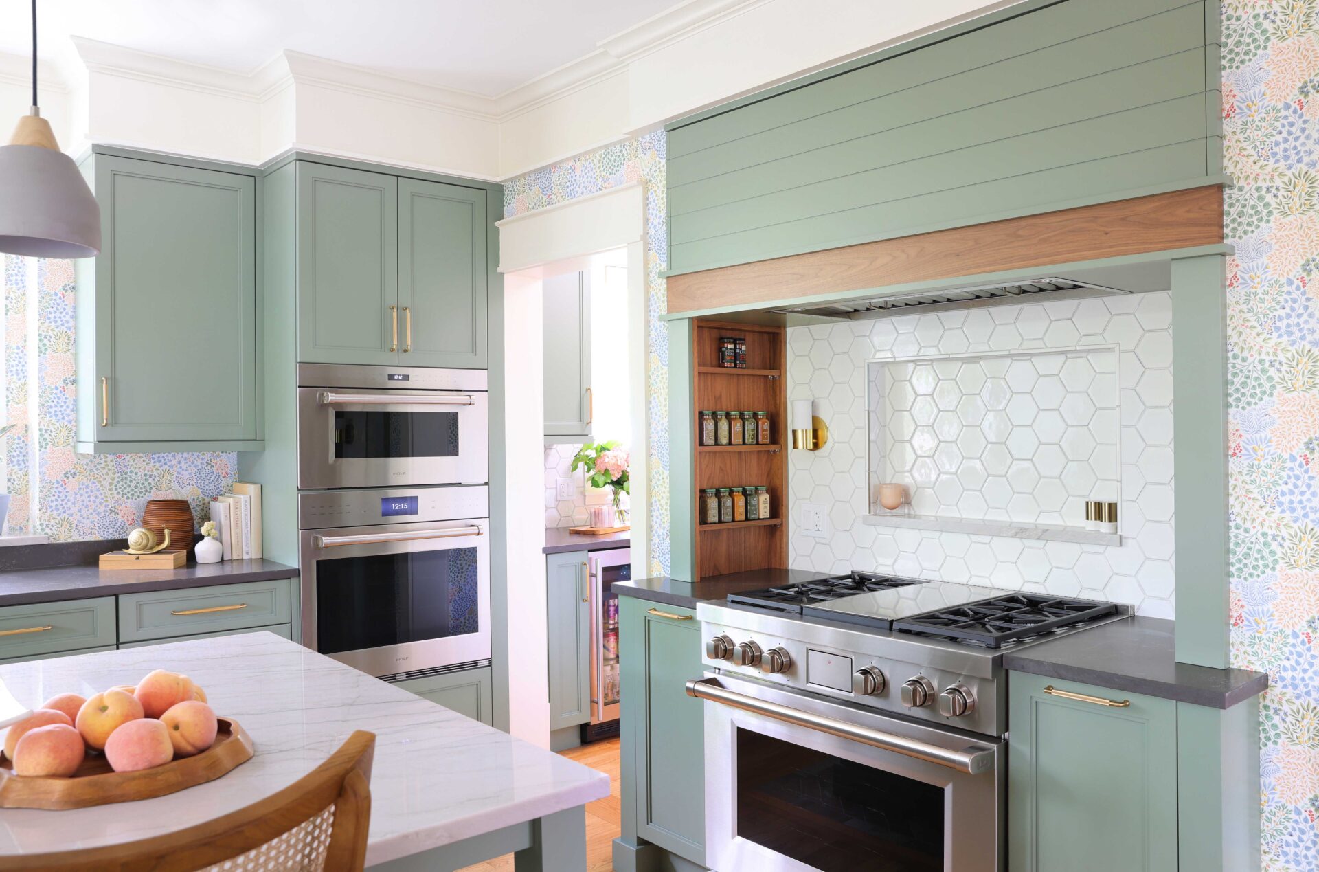 Natural stone white countertop at kitchen island, stacked stainless steel oven and microwave, light green cabinets, green hood with walnut wood trim, white geometric hexagonal backsplash with backsplash shelf and sconces on either side in range alcove with inset walnut spice shelves, stainless steel range