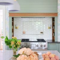 Green range hood with walnut wood trim, white geometric hexagonal tile backsplash with backsplash shelf, sconces, inset walnut spice shelves in range alcove