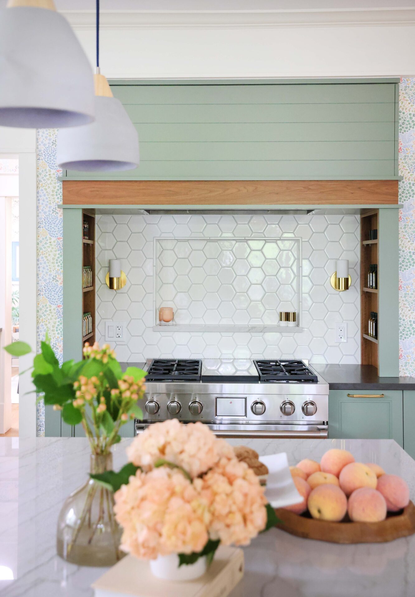 Green range hood with walnut wood trim, white geometric hexagonal tile backsplash with backsplash shelf, sconces, inset walnut spice shelves in range alcove