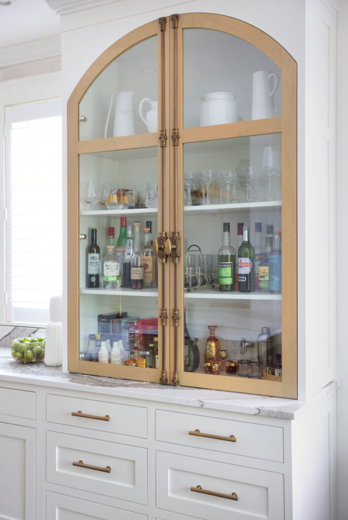 An integrated liquor cabinet including arched doors with clear glass panels and vintage-inspired door pulls.