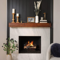 White and grab slab fireplace front with medium stain wood trim, dark blue bead board wainscot paneling accent behind. Mantle holds various candles, vases and books. On the hearth is a vase holding green and dark purple thistle