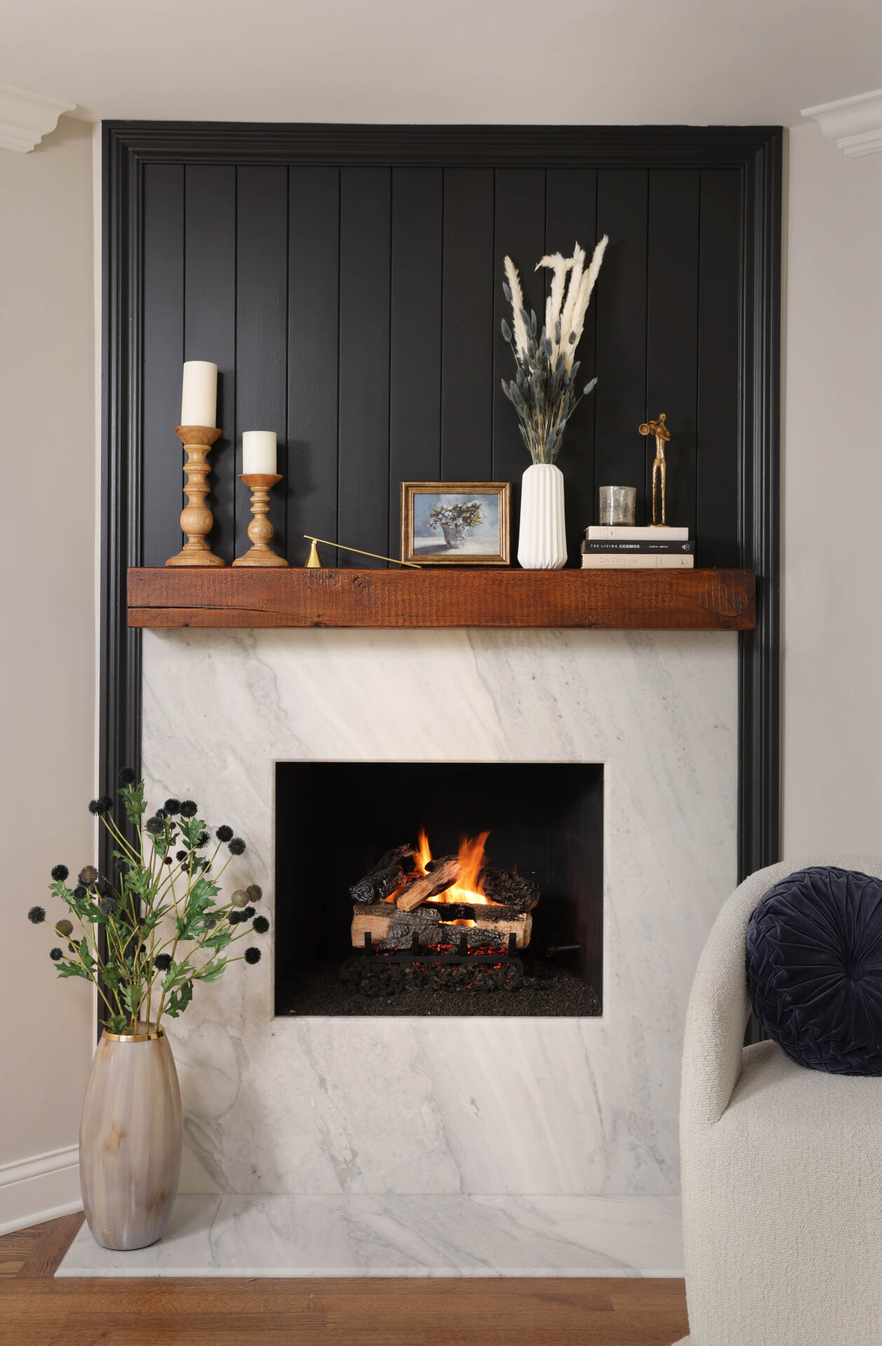 White and grab slab fireplace front with medium stain wood trim wood mantle, dark blue bead board wainscot paneling accent behind. Mantle holds various candles, vases and books. On the hearth is a vase holding green and dark purple thistle