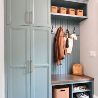 Mudroom with rainforest green cabinets, wood stained mudroom bench with bead board detailing above, and hooks holding a brown jacket and white bag with umbrella and white flowers. Shelving above and below bench. Lower shelves hold shoes and a brown storage bin. Upper shelf holds four small brown storage bins. Herringbone gray tile floor tiles with white grout