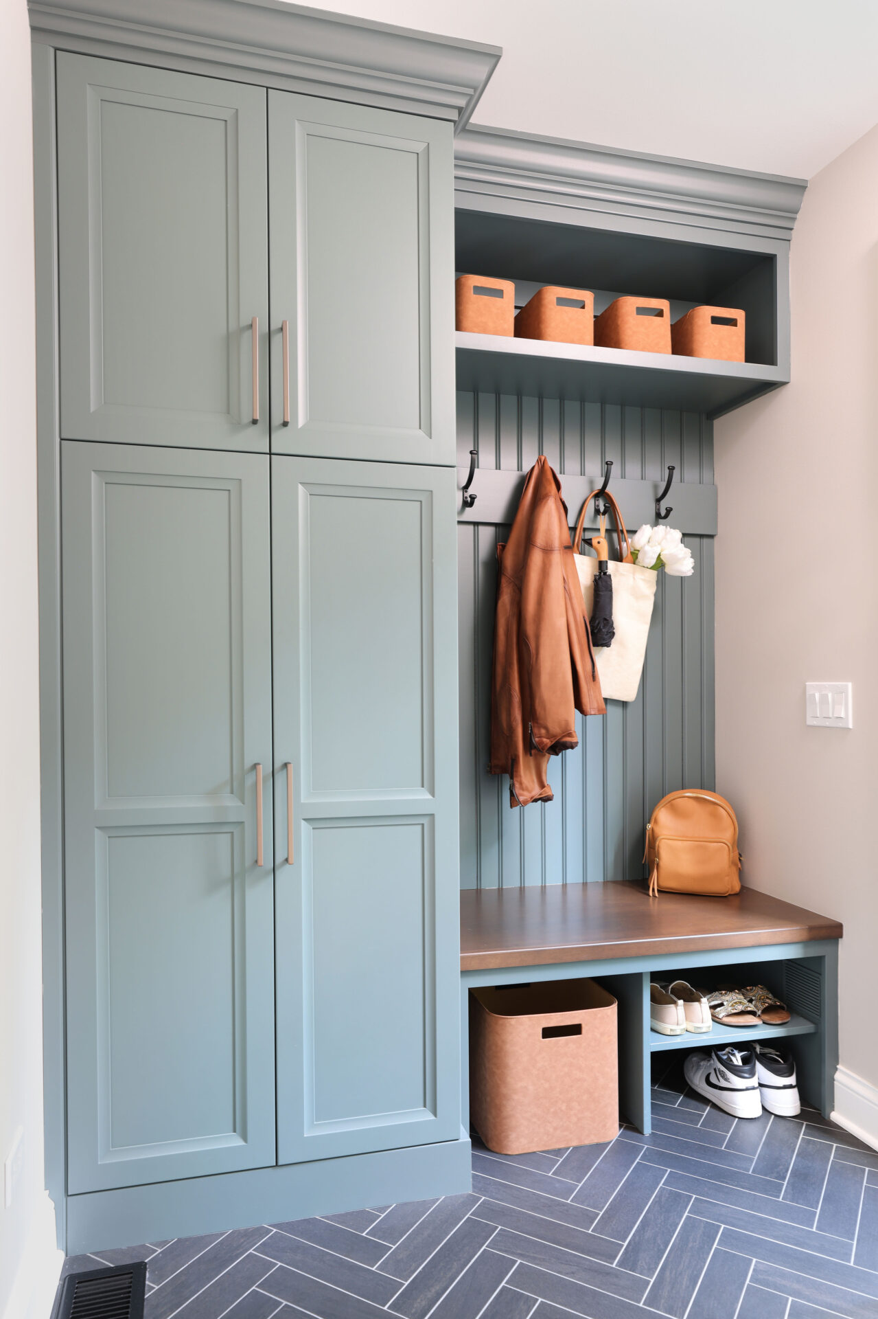 Mudroom with rainforest green cabinets, wood stained mudroom bench with bead board wainscot paneling above, and hooks holding a brown jacket and white bag with umbrella and white flowers. Shelving above and below bench. Lower shelves hold shoes and a brown storage bin. Upper shelf holds four small brown storage bins.