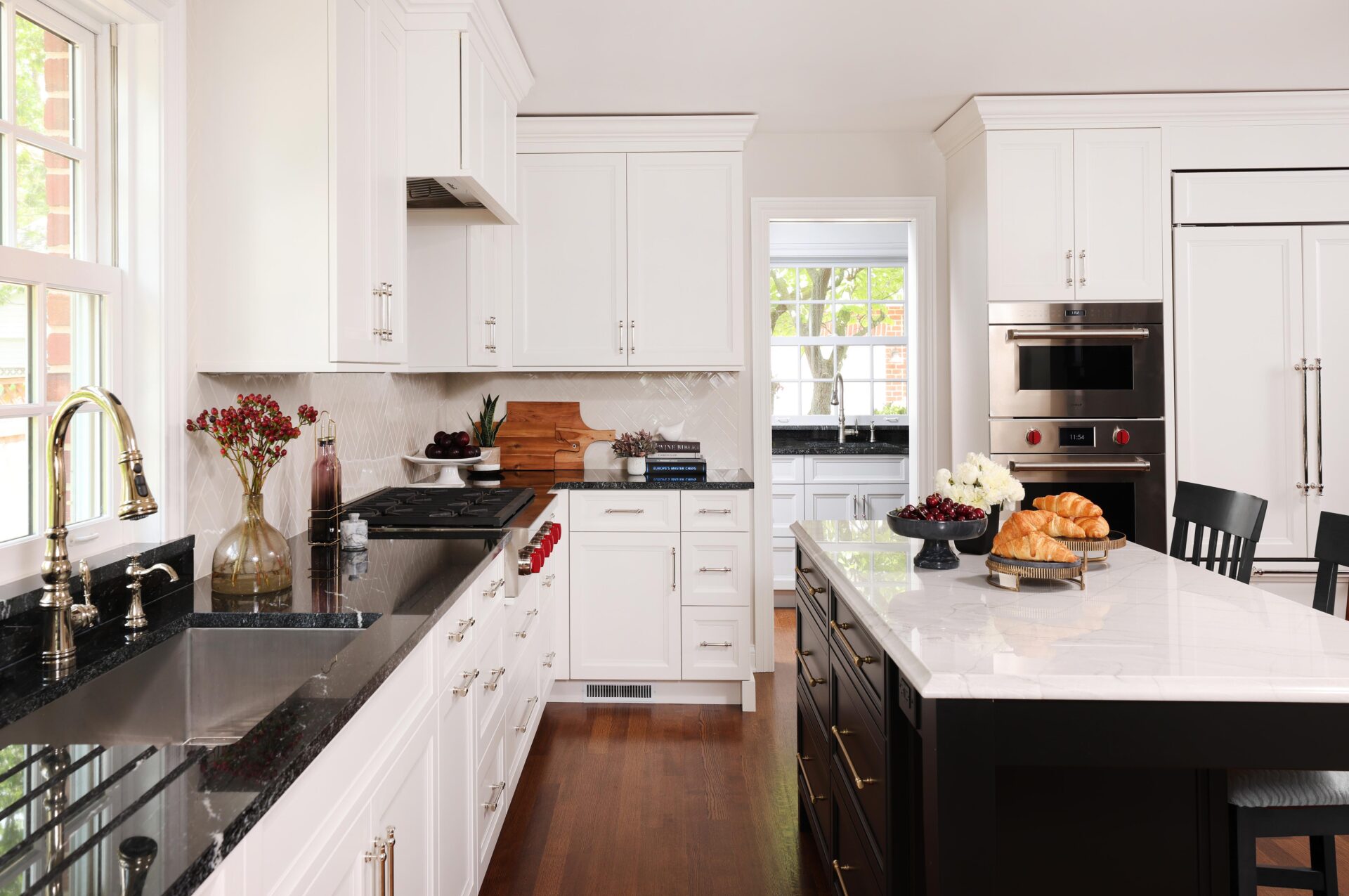 Modern kitchen design featuring white shaker cabinets, black countertops on left perimeter, a large island with black base and white countertop with gold hardware, stainless steel appliances, white paneled refrigerator natural wood flooring. A doorway peeks into a walk-in pantry with sink.