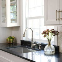 Polished nickel kitchen faucet and cabinet hardware, white kitchen cabinets, black countertops, one upper cabinet glass-fronted kitchen cabinet