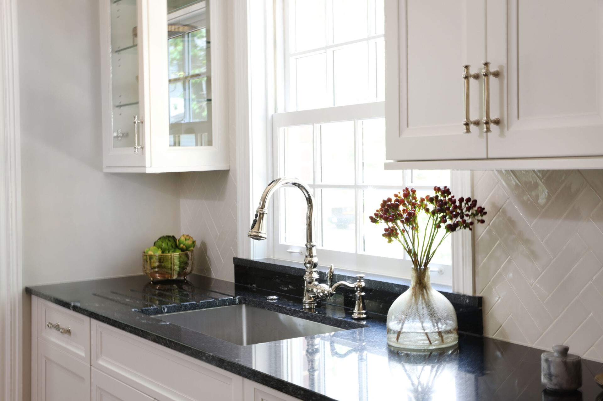 Polished nickel kitchen faucet and cabinet hardware, white kitchen cabinets, black countertops, one upper cabinet glass-fronted kitchen cabinet