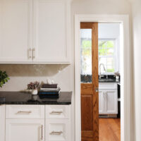 White kitchen cabinets with black countertops, wood pocket door half open showing a walk-in pantry with white cabinets, black countertops and sink with window above