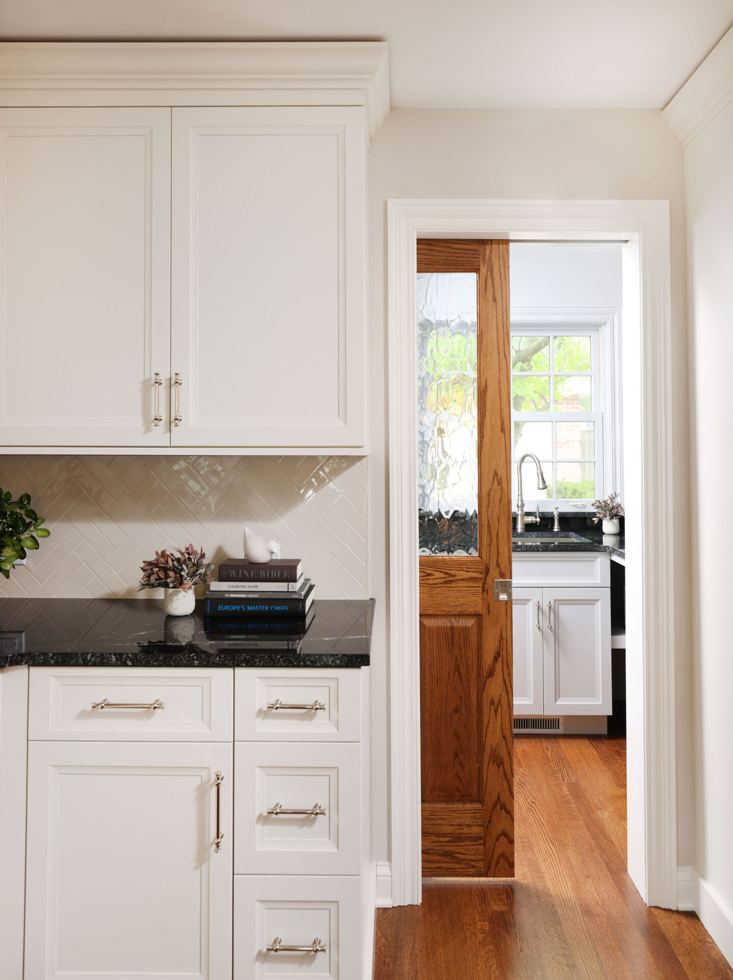 White kitchen cabinets with black countertops, wood pocket door half open showing a walk-in pantry with white cabinets, black countertops and sink with window above