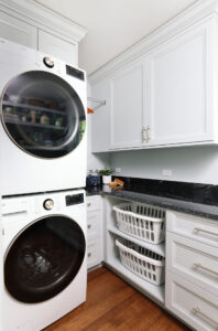 White stacked washer and dryer with black circular doors surrounded by white cabinetry, black countertops. Below the counter is a set of shelves holding two white laundry baskets