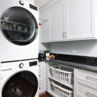 White stacked washer and dryer with black circular doors surrounded by white cabinetry, black countertops. Below the counter is a set of shelves holding two white laundry baskets