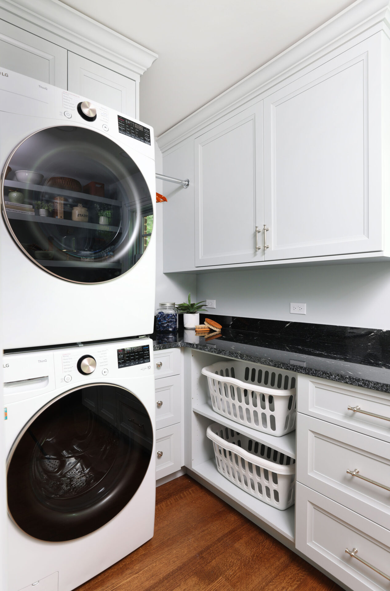White stacked washer and dryer with black circular doors surrounded by white cabinetry, black countertops. Below the counter is a set of shelves holding two white laundry baskets