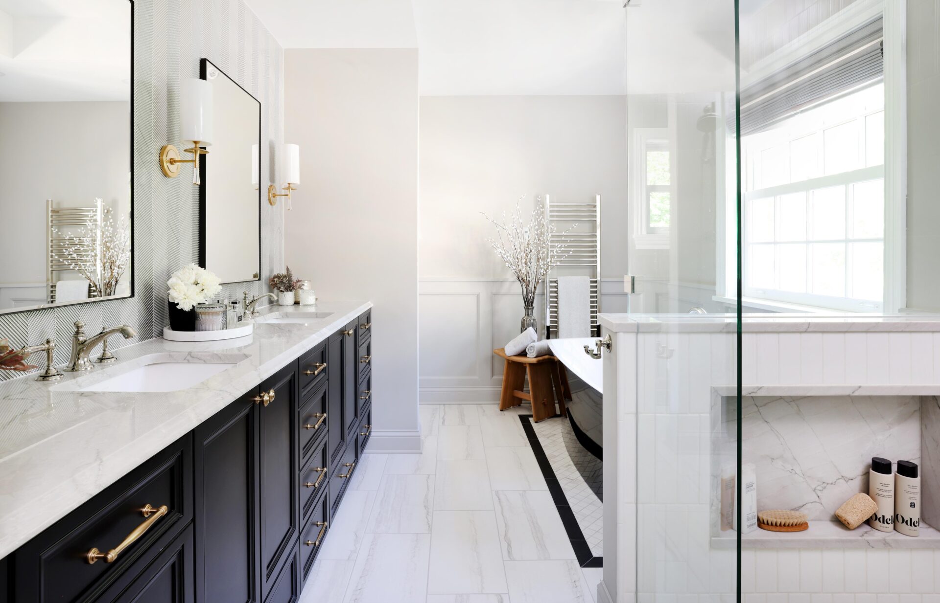 Double black vanity with quartzite white countertops on the left with two black-rimmed mirror above the two sinks and gold sconces with white lights. On the right is a black freestanding tub with white interior in the background near a stainless steel towel warmer. On the right in the foreground is the edge of a glass-enclosed shower with a white tile half wall and shower niche holding shampoo, conditioner and loofahs