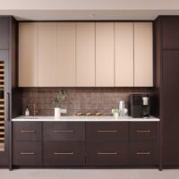 The modern beverage center features rift cut white oak cabinetry, ceramic tile backsplash, quartz countertops, wine fridge, integrated refrigerator drawers, brushed brass elements, and fireclay basin.