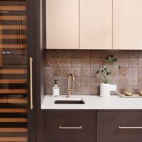 The modern beverage center features rift cut white oak cabinetry, ceramic tile backsplash, quartz countertops, wine fridge, integrated refrigerator drawers, brushed brass elements, and fireclay basin.