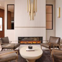 Modern minimalist fireplace features vertical ceramic tile surround and brushed brass decorative band. Striking brass light fixture hangs overhead, complimenting the verticality of the design. The electric fireplace itself delivers warm ambience without the need for venting.