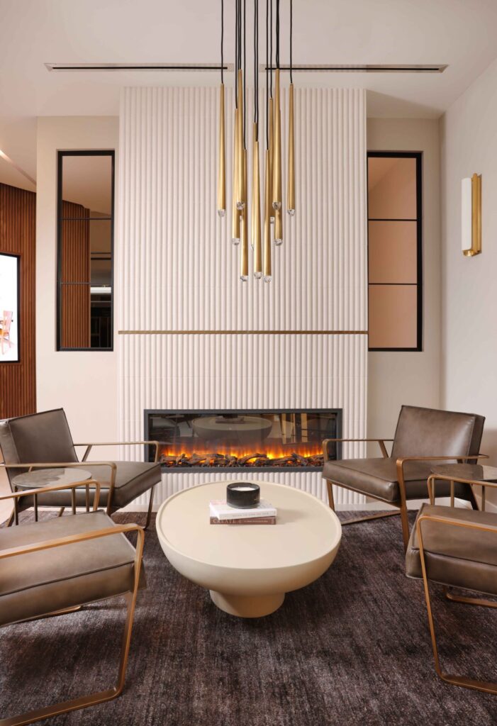 Modern minimalist fireplace features vertical ceramic tile surround and brushed brass decorative band. Striking brass light fixture hangs overhead, complimenting the verticality of the design. The electric fireplace itself delivers warm ambience without the need for venting.