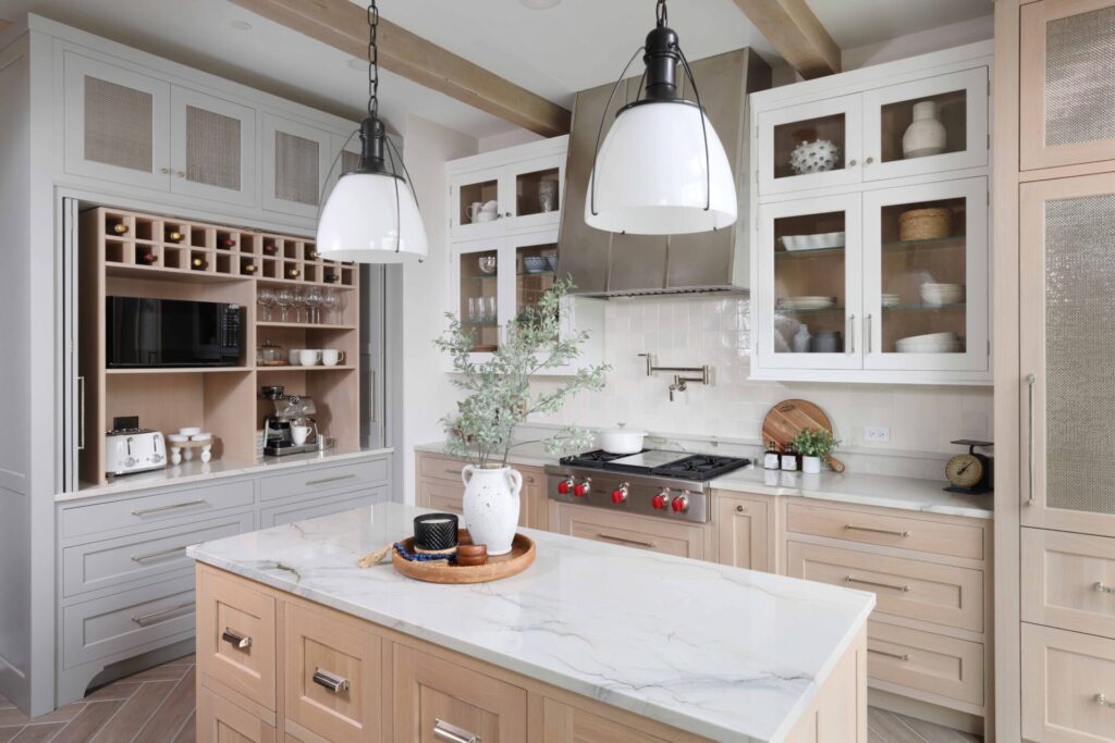 Modern farmhouse kitchen includes rift cut white oak cabinets in natural, white and gray painted finishes, custom hutch with decorative feet and bi-fold pocket doors, monochromatic backsplash tile, island with baking center, natural stone countertops, polished nickel pot filler, stainless steel range hood, black pendant lights and porcelain floor tile in a herringbone pattern.