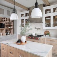 Modern farmhouse kitchen includes rift cut white oak cabinets in natural, white and gray painted finishes, custom hutch with decorative feet and bi-fold pocket doors, monochromatic backsplash tile, island with baking center, natural stone countertops, polished nickel pot filler, stainless steel range hood, black pendant lights and porcelain floor tile in a herringbone pattern.