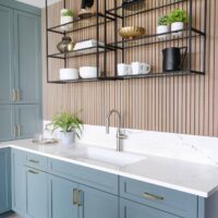 Above the spacious low-divide sink, matte-black metal shelves replace upper cabinets and offer more bold shapes, creating layered contrast in front of the oak slat paneling. Vertical wood slats at the sink wall and island are a rift cut white oak wood species that brings out beautiful color variations in stains.