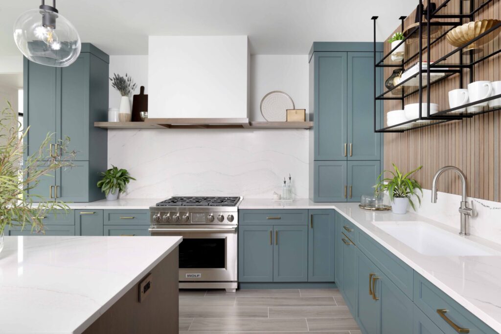 The color forward contemporary kitchen features classic shaker cabinetry; a stylish rainforest blue-green cabinetry finish; paneled appliances, such as the refrigerator, blend with surrounding cabinetry and reinforce this aesthetic, allowing the cabinetry color to take the spotlight while creating a fluid geometric presence. Also visible is a minimalist range hood; custom drywall, with a subtle wood trim, anchors the range wall, and provides unique shelving on either end. The hood is finished to blend seamlessly into the surrounding walls rather than competing for attention. Quartz countertops and full-height backsplash with soft veining further balance the space with a calm, neutral surface.