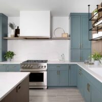 The color forward contemporary kitchen features classic shaker cabinetry; a stylish rainforest blue-green cabinetry finish; paneled appliances, such as the refrigerator, blend with surrounding cabinetry and reinforce this aesthetic, allowing the cabinetry color to take the spotlight while creating a fluid geometric presence. Also visible is a minimalist range hood; custom drywall, with a subtle wood trim, anchors the range wall, and provides unique shelving on either end. The hood is finished to blend seamlessly into the surrounding walls rather than competing for attention. Quartz countertops and full-height backsplash with soft veining further balance the space with a calm, neutral surface.