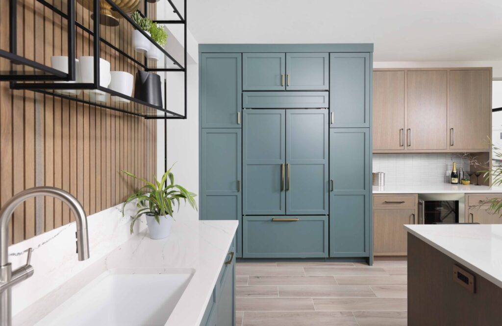 The color forward contemporary kitchen features classic shaker cabinetry in a stylish rainforest blue-green finish, paneled appliances, such as the refrigerator, blend with surrounding cabinetry and reinforce this aesthetic, allowing the cabinetry color to take the spotlight while creating a fluid geometric presence. Beverage Center: Classic shaker cabinetry in the kitchen is mixed with sleek slab-front doors at the beverage center — a modern take on inset cabinets, which typically leans more traditional — creating visual interest without overwhelming the space. The beverage center also offers a moment to introduce shine, with a glass tile backsplash that distinguishes the area from the main kitchen, while still tying into the overall palette. A beverage refrigerator and concealed bar pullout keep bottles tucked away when not entertaining.
