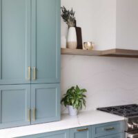The color forward contemporary kitchen features a minimalist range hood; custom drywall with a subtle wood trim anchors the range wall, and provides unique shelving on either end. The hood is finished to blend seamlessly into the surrounding walls rather than competing for attention. Quartz countertops and full-height backsplash with soft veining further balance the space with a calm, neutral surface.