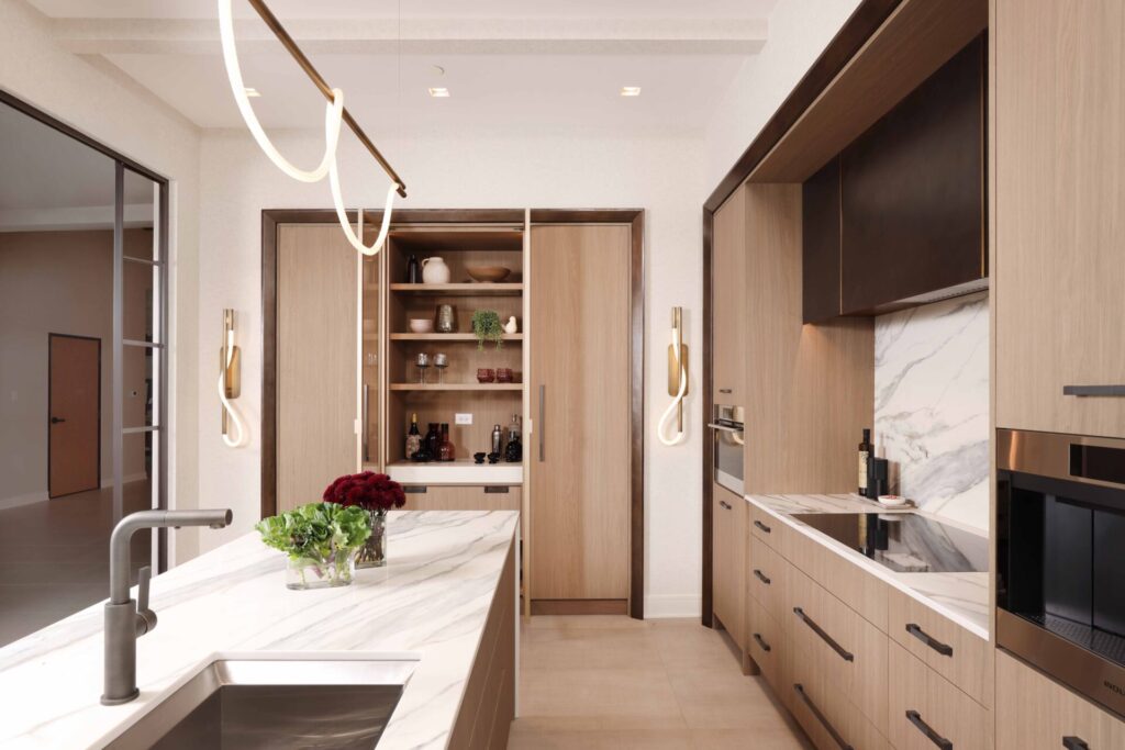 The curated modern kitchen features textured melamine cabinetry; a suite of integrated appliances including steam oven, coffee machine and induction cooktop; blackened steel range hood with brass detailing; manmade quartz waterfall island, backsplash and countertops that mimic the look of marble; loop lighting in the wall sconces and above the island.