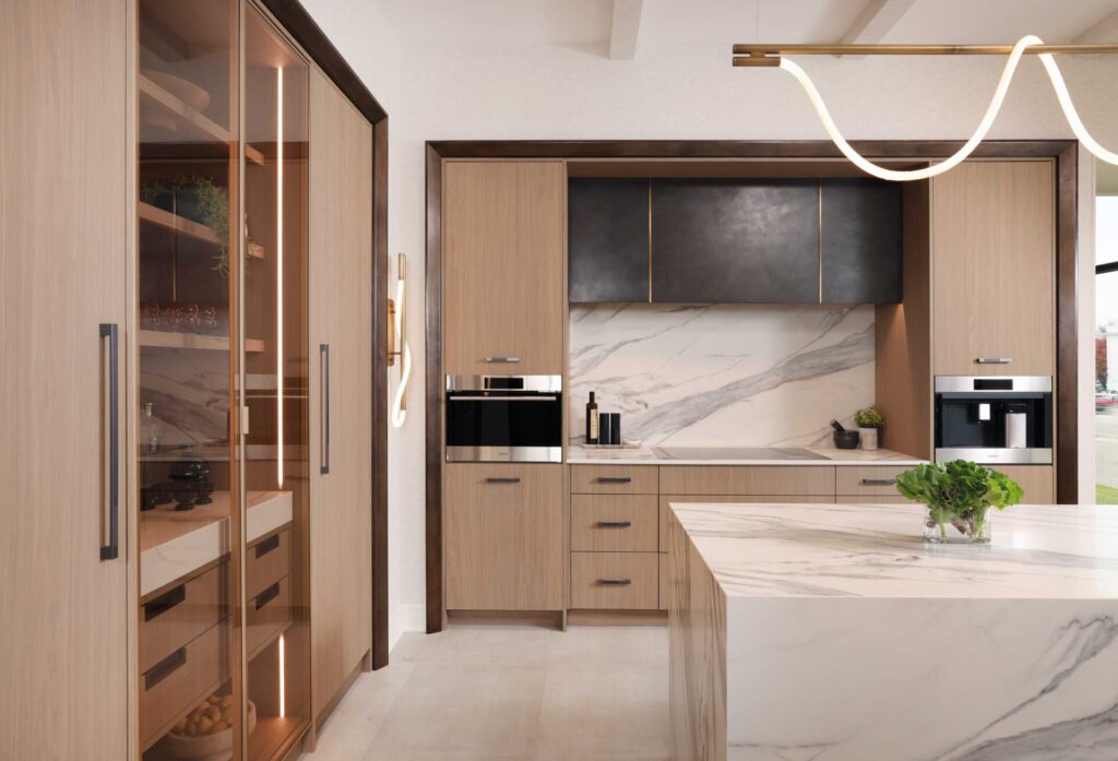 The curated modern kitchen features textured melamine cabinetry; a suite of integrated appliances including steam oven, coffee machine and induction cooktop; blackened steel range hood with brass detailing; manmade quartz waterfall island, backsplash and countertops that mimic the look of marble; loop lighting in the wall sconces and above the island.