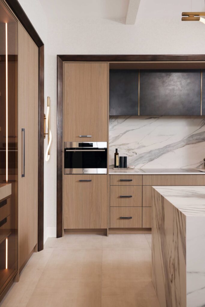 The curated modern kitchen features textured melamine cabinetry; a suite of integrated appliances including steam oven, coffee machine and induction cooktop; blackened steel range hood with brass detailing; manmade quartz waterfall island, backsplash and countertops that mimic the look of marble; loop lighting in the wall sconces and above the island.