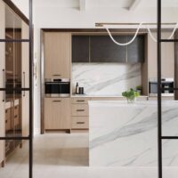 The curated modern kitchen features textured melamine cabinetry; a suite of integrated appliances including steam oven, coffee machine and induction cooktop; blackened steel range hood with brass detailing; manmade quartz waterfall island, backsplash and countertops that mimic the look of marble; loop lighting in the wall sconces and above the island.