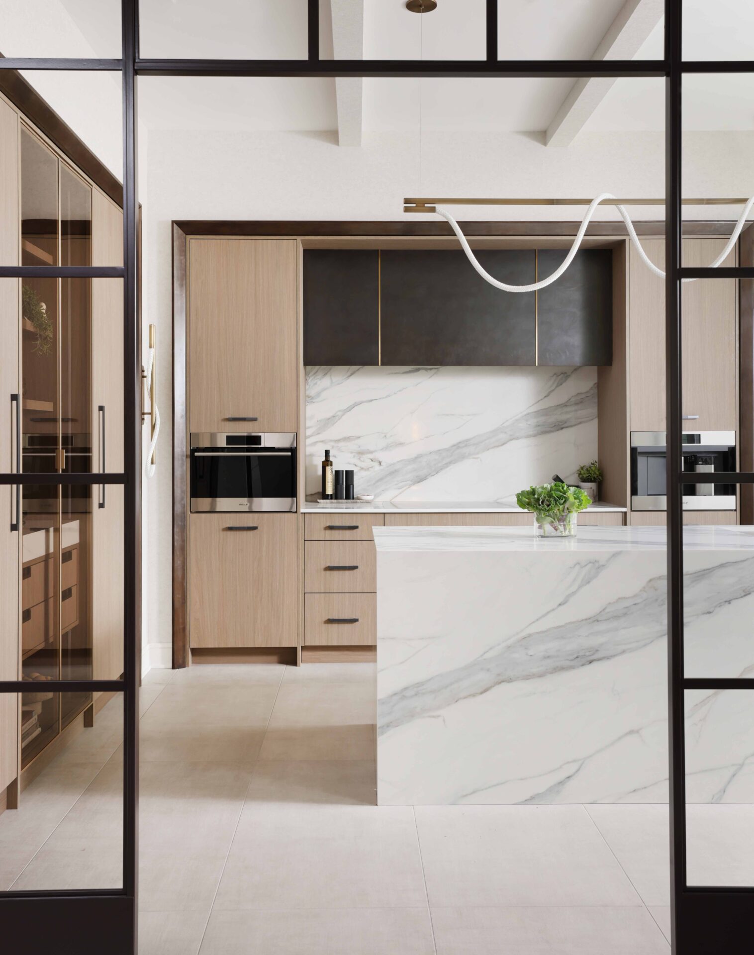 The curated modern kitchen features textured melamine cabinetry; a suite of integrated appliances including steam oven, coffee machine and induction cooktop; blackened steel range hood with brass detailing; manmade quartz waterfall island, backsplash and countertops that mimic the look of marble; loop lighting in the wall sconces and above the island.