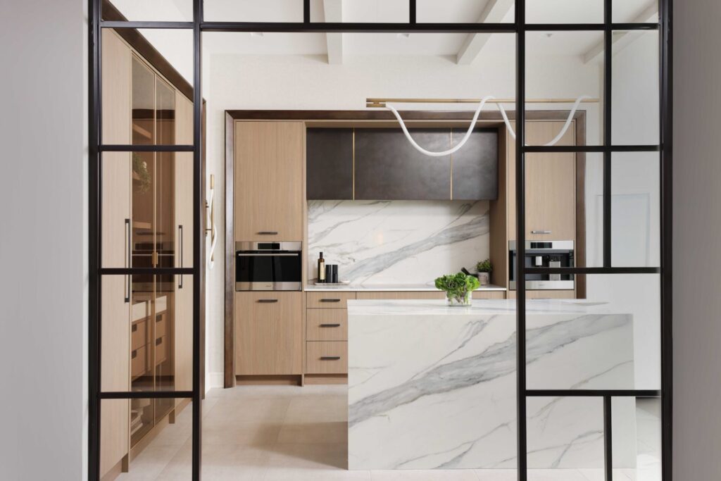 The curated modern kitchen features textured melamine cabinetry; a suite of integrated appliances including steam oven, coffee machine and induction cooktop; blackened steel range hood with brass detailing; manmade quartz waterfall island, backsplash and countertops that mimic the look of marble; loop lighting in the wall sconces and above the island.