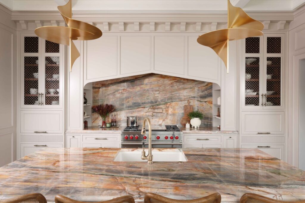 The elevated new traditional kitchen features painted cabinets throughout, color inspired by veining in backsplash and counters. Range area has recessed spice niches; flanking cabinets feature walnut interiors, ornamentation, glass shelves, accent lighting. Full height quartzite backsplash with rose and caramel veining. 48-inch range with Tudor style arched hood. Brushed bronze pot filler. Cherry stained island, thick quartzite counter with double ogee edge, furniture style legs and ample seating; sink with faucet in brushed bronze. Luxe gold pendant lights.