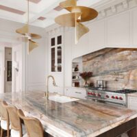 The elevated new traditional kitchen features painted cabinets throughout, color inspired by veining in backsplash and counters. Range area has recessed spice niches; flanking cabinets feature walnut interiors, ornamentation, glass shelves, accent lighting. Full height quartzite backsplash with rose and caramel veining. 48-inch range with Tudor style arched hood. Brushed bronze pot filler. Cherry stained island, thick quartzite counter with double ogee edge, furniture style legs and ample seating; sink with faucet in brushed bronze. Luxe gold pendant lights.