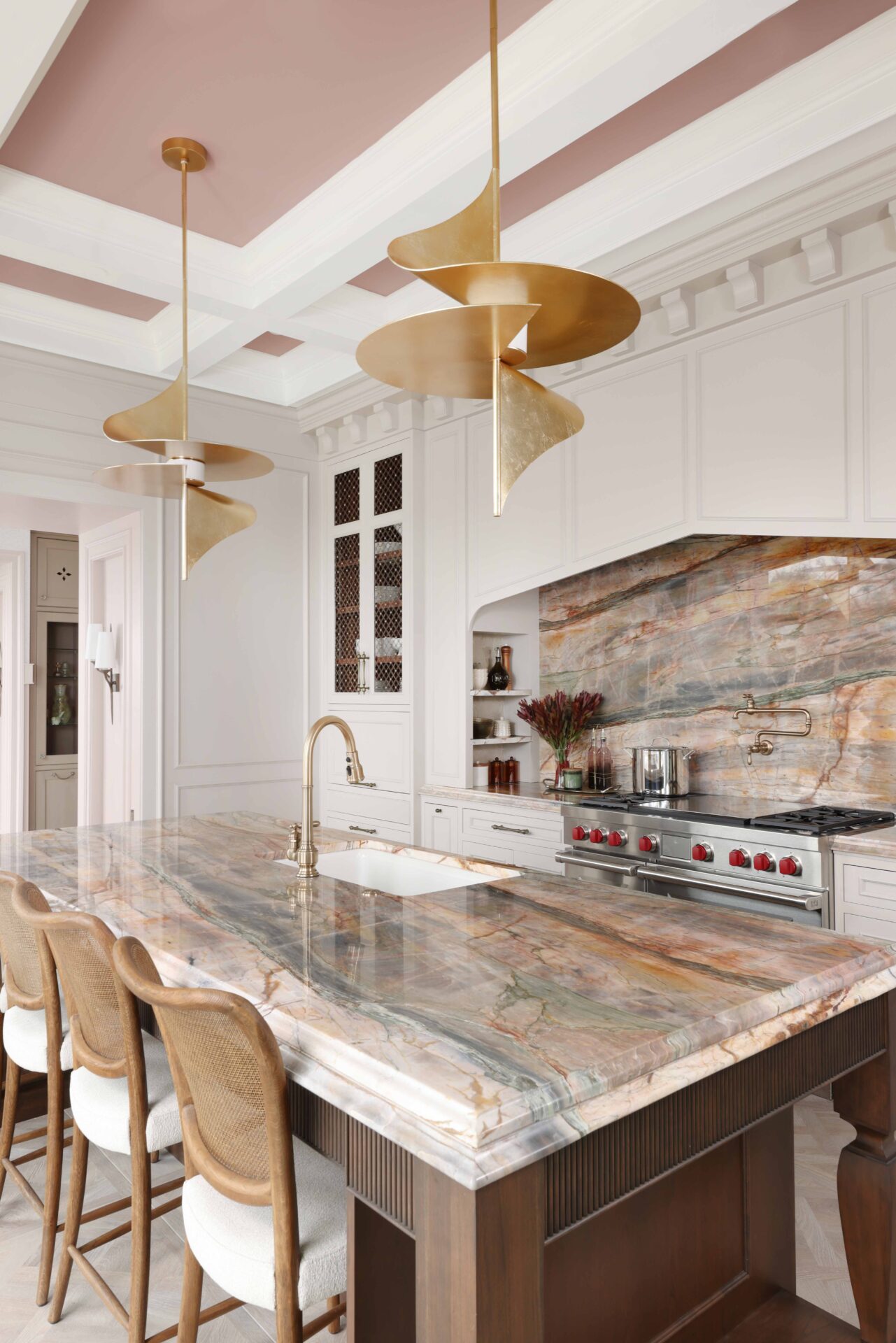 The elevated new traditional kitchen features painted cabinets throughout, color inspired by veining in backsplash and counters. Range area has recessed spice niches; flanking cabinets feature walnut interiors, ornamentation, glass shelves, accent lighting. Full height quartzite backsplash with rose and caramel veining. 48-inch range with Tudor style arched hood. Brushed bronze pot filler. Cherry stained island, thick quartzite counter with double ogee edge, furniture style legs and ample seating; sink with faucet in brushed bronze. Luxe gold pendant lights.