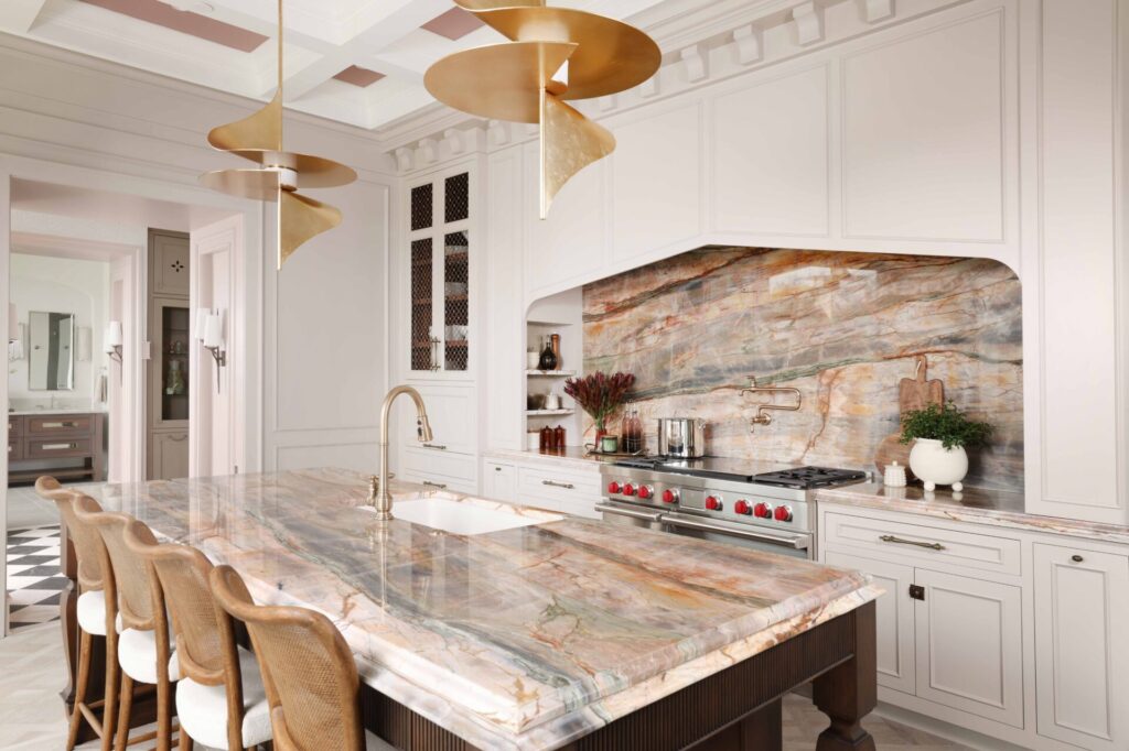 The elevated new traditional kitchen features painted cabinets throughout, color inspired by veining in backsplash and counters. Range area has recessed spice niches; flanking cabinets feature walnut interiors, ornamentation, glass shelves, accent lighting. Full height quartzite backsplash with rose and caramel veining. 48-inch range with Tudor style arched hood. Brushed bronze pot filler. Cherry stained island, thick quartzite counter with double ogee edge, furniture style legs and ample seating; sink with faucet in brushed bronze. Luxe gold pendant lights.