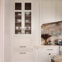 The elevated new traditional kitchen features painted cabinets, color inspired by veining in backsplash and counters. Cabinet interiors finished in walnut; accessories include a knife block drawer and lift open mechanisms. Flanking cabinets feature ornamentation, glass shelves, accent lighting. Elegant moldings, tall baseboards, corbels, tray style coffered ceiling add layers of architectural detail.
