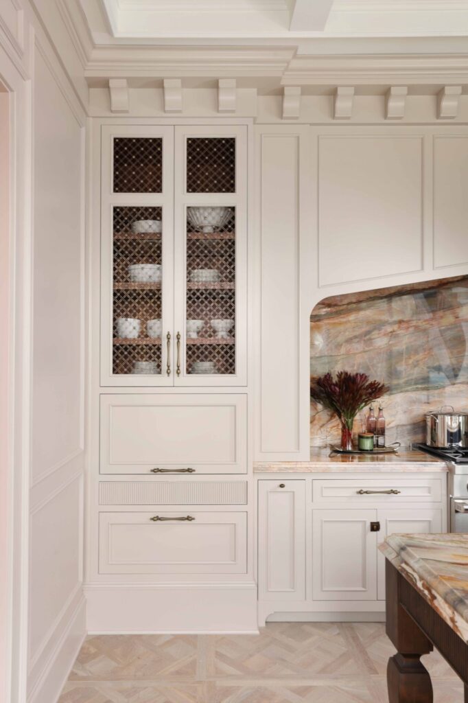 The elevated new traditional kitchen features painted cabinets, color inspired by veining in backsplash and counters. Cabinet interiors finished in walnut; accessories include a knife block drawer and lift open mechanisms. Flanking cabinets feature ornamentation, glass shelves, accent lighting. Elegant moldings, tall baseboards, corbels, tray style coffered ceiling add layers of architectural detail.